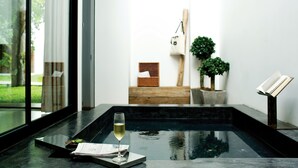 A modern spa area with a glass-enclosed hot tub, a glass of white wine, and a small table with a book and a menu. - The Library (Koh Samui)