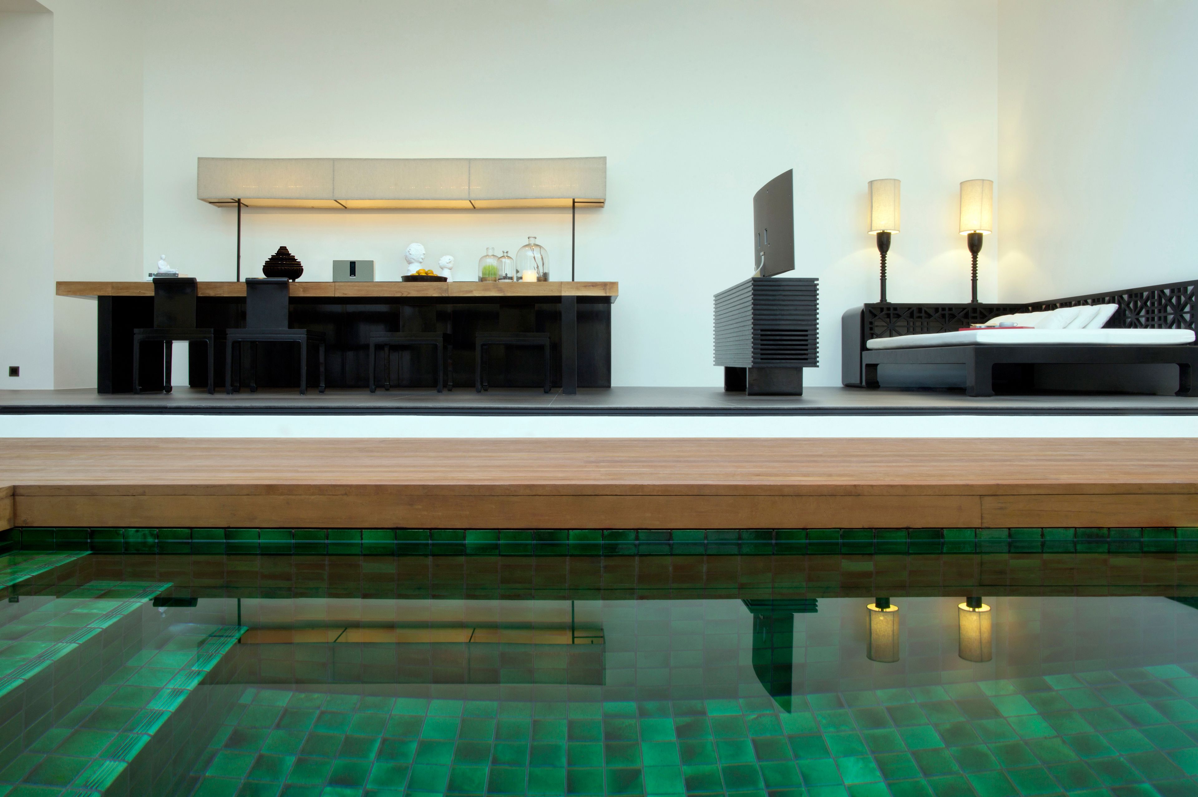 A modern hotel room with a wooden reception desk, a sofa, a TV, and a pool with green tiles.