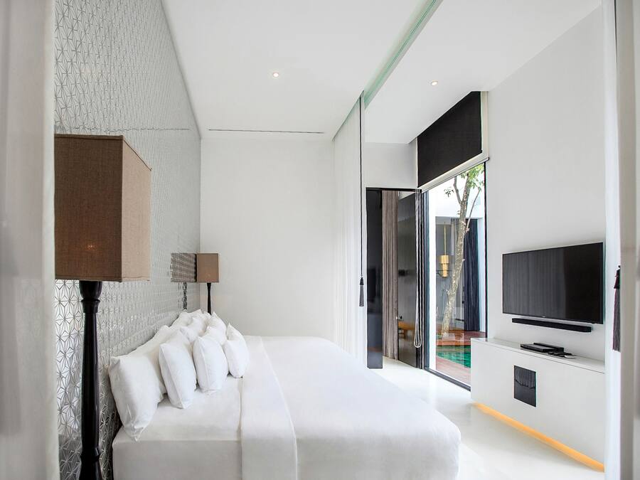 A modern bedroom with a large bed, a flat-screen TV mounted on the wall, and a view of the outdoors through a glass door.