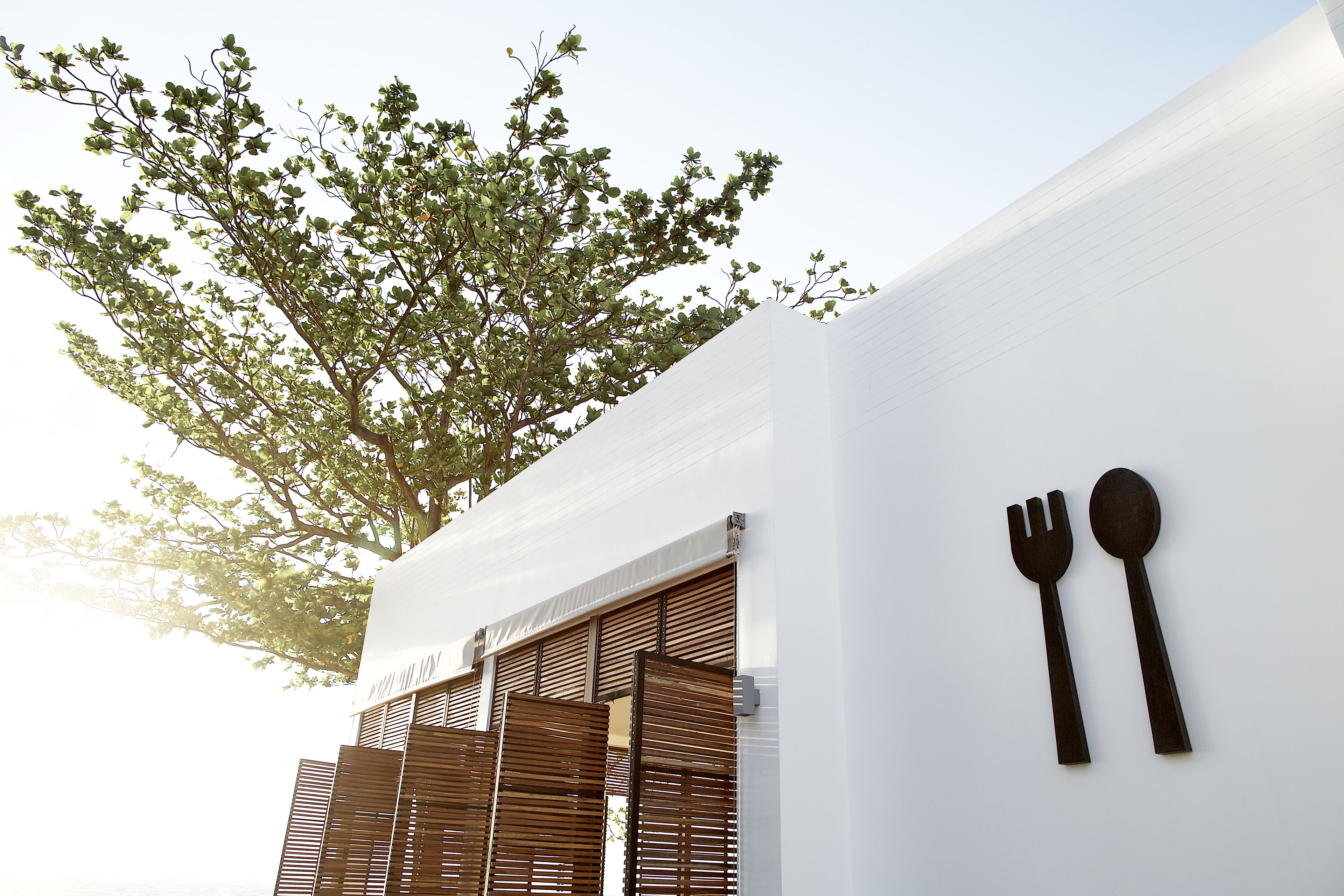A white building with a tree and wooden shutters, and a wall-mounted symbol of a fork and spoon.