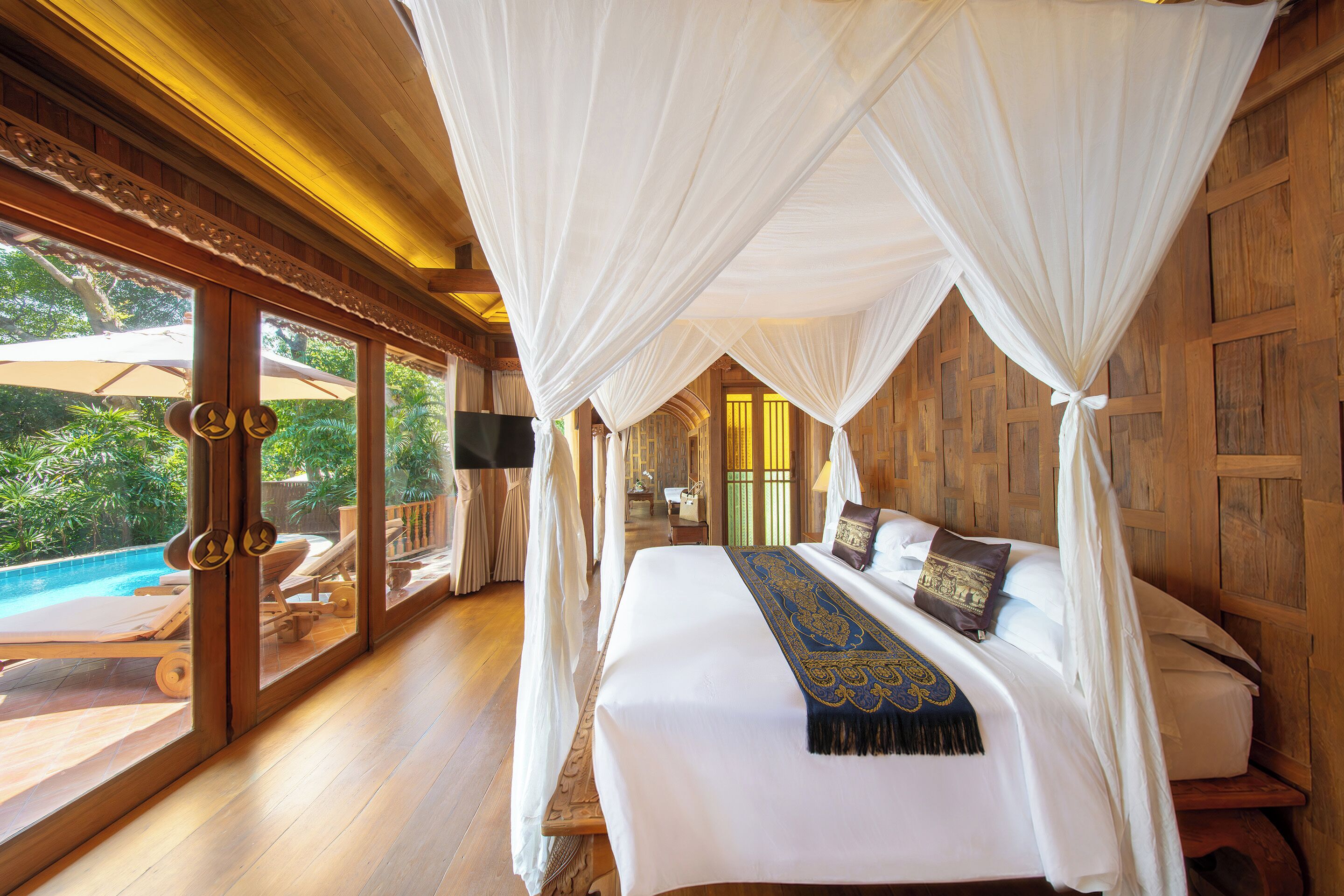 A bedroom with a large bed, white curtains, and a view of a pool and greenery outside.