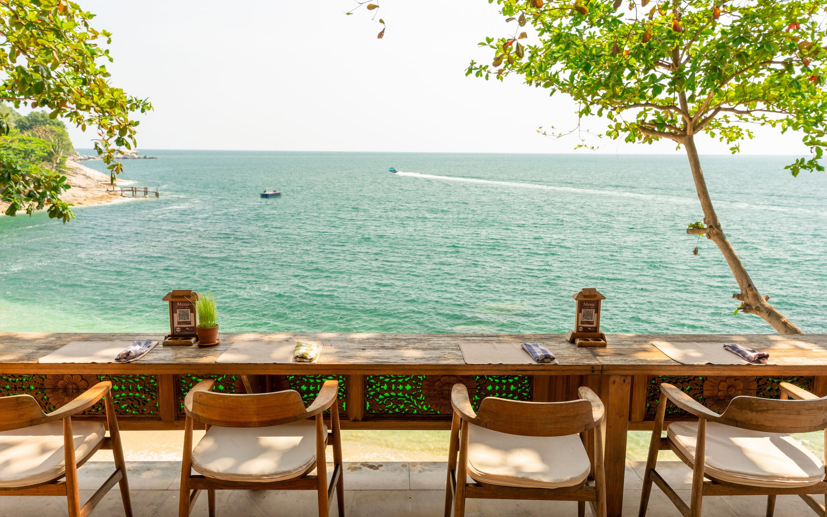 Une table en bois, entourée de chaises, est placée face à la mer. On aperçoit un bateau au loin et un arbre sur la droite.