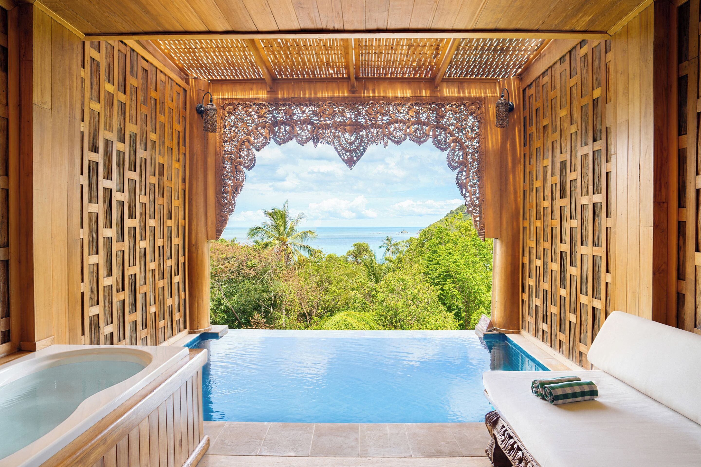 A wooden spa area with a hot tub and a lounge chair, overlooking a pool and a tropical landscape.