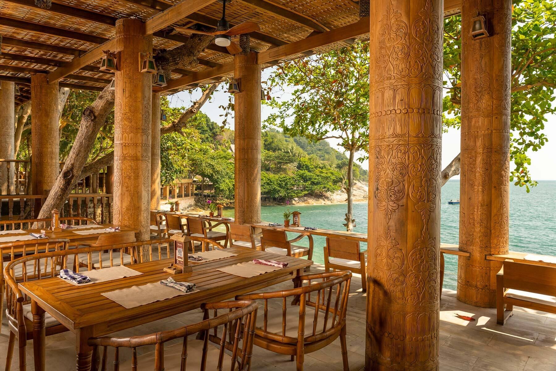 An outdoor dining area with wooden tables and chairs, surrounded by intricately carved columns, overlooking a beach and the sea.