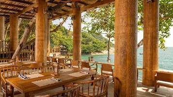 An outdoor dining area with wooden tables and chairs, surrounded by intricately carved columns, overlooking a beach and the sea.