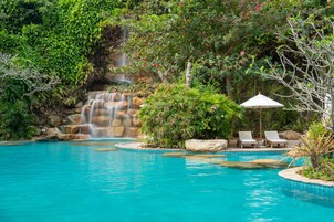 Una piscina con cascada, rodeada de exuberante vegetación y un área de descanso con sillas y una sombrilla.