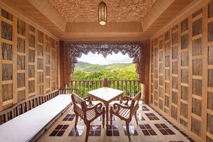 Un balcón de madera con una mesa y sillas, vistas a colinas verdes y un techo decorativo.