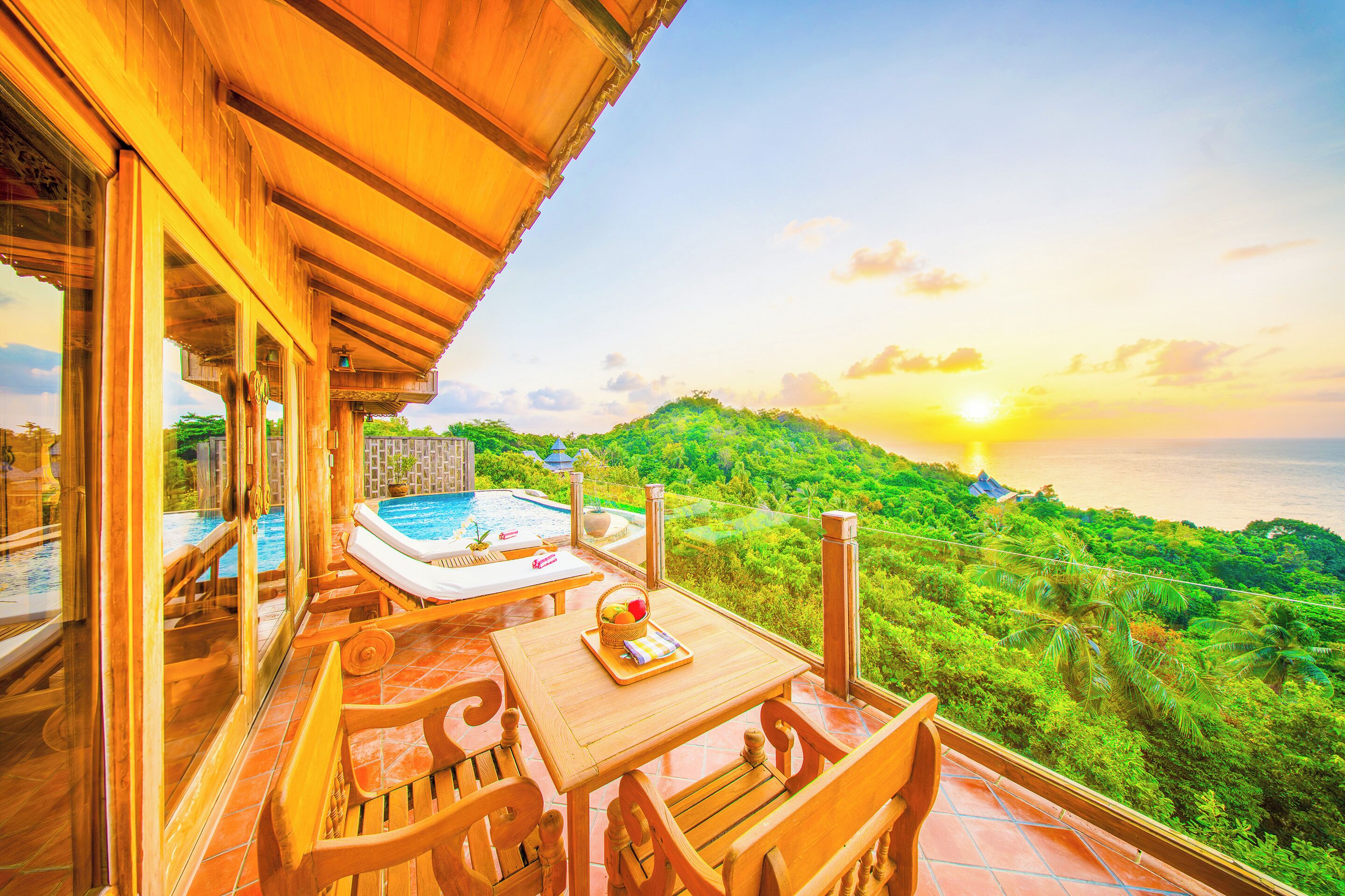 A wooden deck with a pool, a table, and chairs overlooking a lush landscape and the sea at sunset.