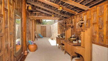 Interior rústico con paredes de madera, pared de piedra y techo de madera. Se observa un espejo, una olla grande y una mesa con varios objetos.