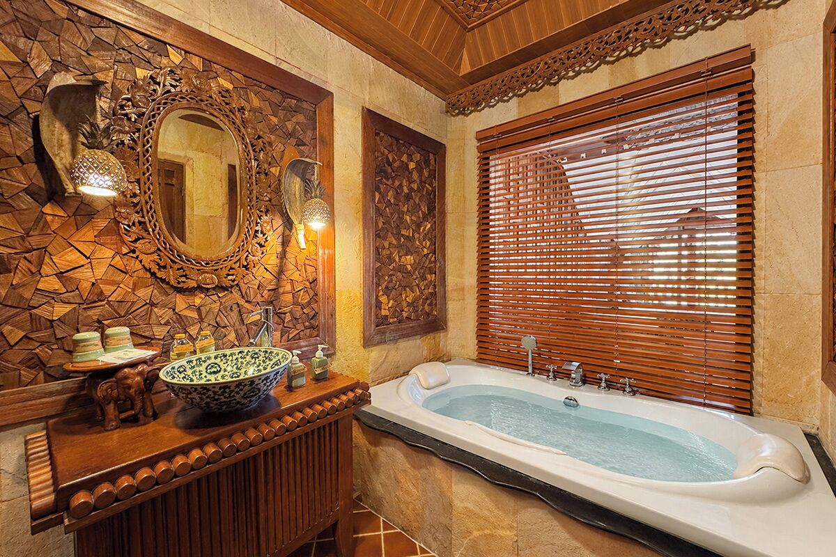 A bathroom with a stone wall, wooden vanity, a large bathtub, and a window with blinds.