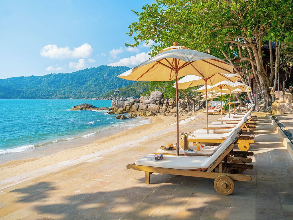 Un complexe hôtelier situé en bord de mer, doté de chaises longues et de parasols, offrant une vue dégagée sur la mer et bordé d’une côte rocheuse.