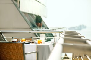 A table set with breakfast items, including a glass of orange juice, a cup, and a plate with food, on a balcony with a railing and a view of the sea.