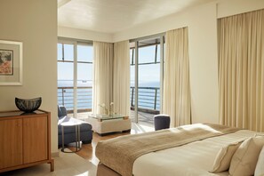 Une chambre d’hôtel avec un lit, un coin salon avec une table, une vue sur la mer et un balcon avec des rideaux.