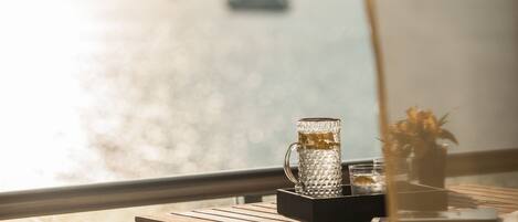 Una mesa con una jarrón de vidrio y un florero con flores, con vistas a un cuerpo de agua donde se distingue un barco a lo lejos.