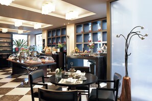 A hotel dining area with a checkered floor, round tables, and chairs. There are glass display cases with food items and a decorative plant. - Daios Luxury Living (Thessaloniki)