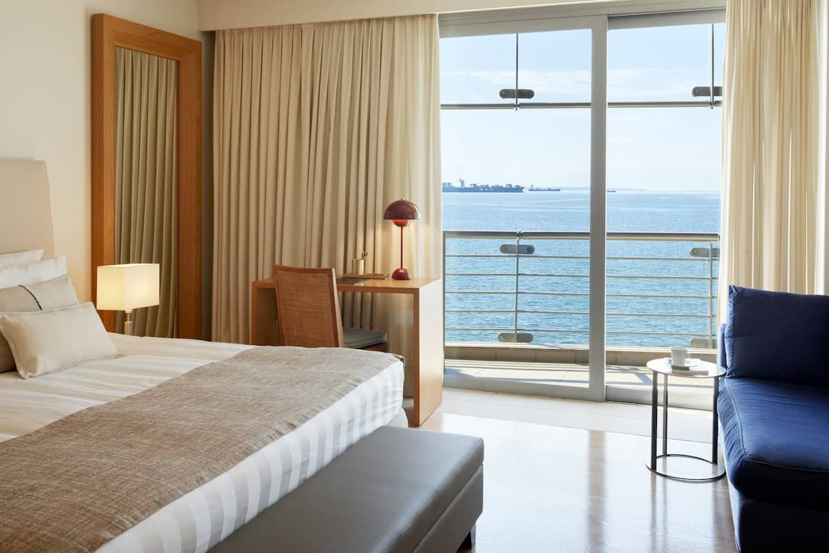 Een hotelkamer met een bed, een bureau, een stoel, een blauwe bank, een lamp en uitzicht op zee.