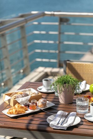 Una mesa dispuesta con pasteles, salsas y una planta en maceta en una terraza con barandilla y vista al mar.