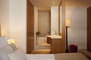 Una habitación de hotel con cama, baño con lavamanos y espejo, y una escalera que lleva a la zona de la ducha.