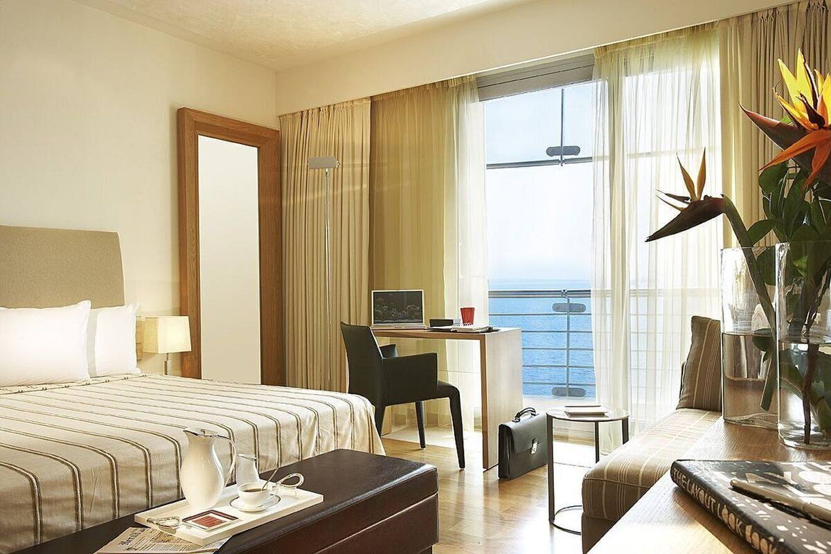 Een hotelkamer met een bed, een bureau met computer, een stoel, een kleintje tafeltje, een koffer en uitzicht op zee.