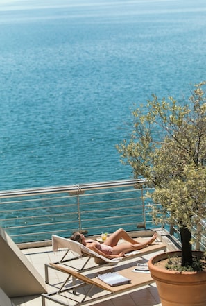 Un balcon avec vue sur la mer, un fauteuil de relaxation, une plante en pot et une petite table avec une boisson.