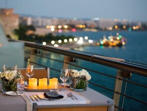 A table set for dining with candles, glasses, and flowers, overlooking a waterfront with city lights in the background.