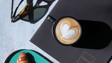 Une tasse de café noir décorée d’un motif en latte art, une paire de lunettes de soleil et deux croissants sur une assiette de couleur turquoise.