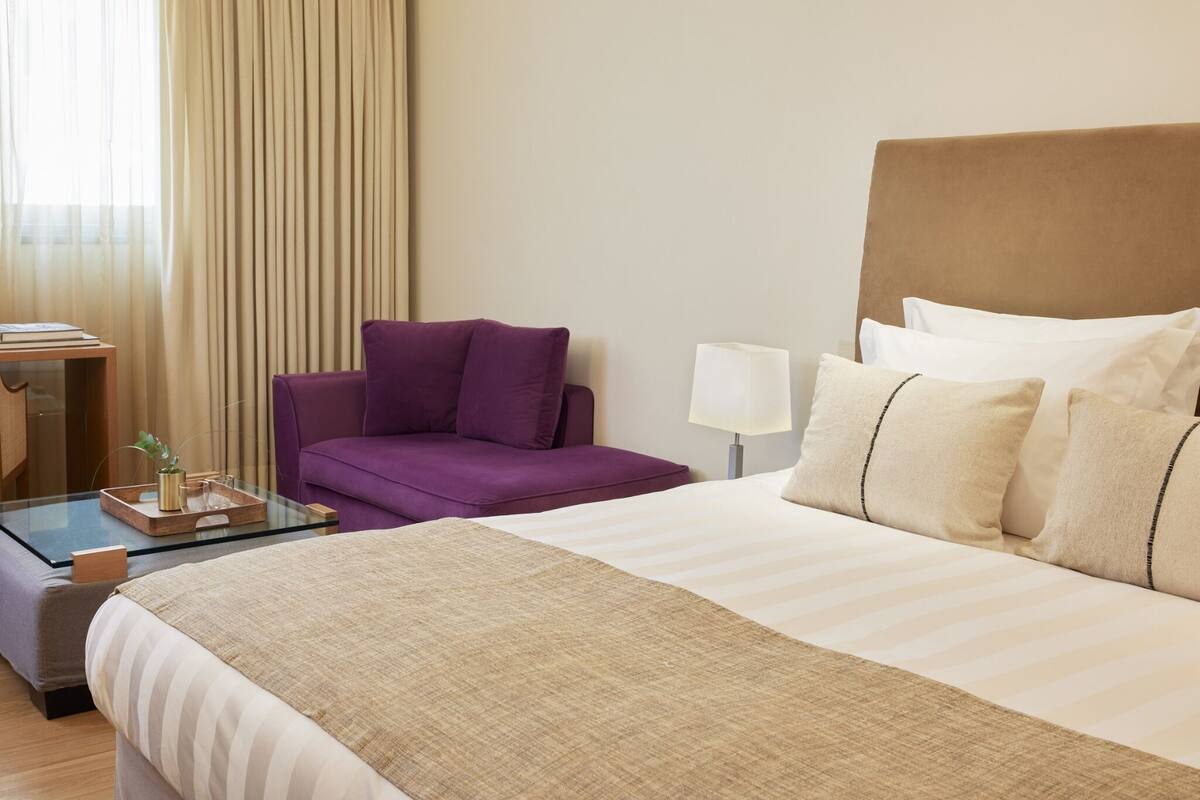 Een hotelkamer met een bed, een paarse fauteuil, een tafeltje met een vaas en een lamp.