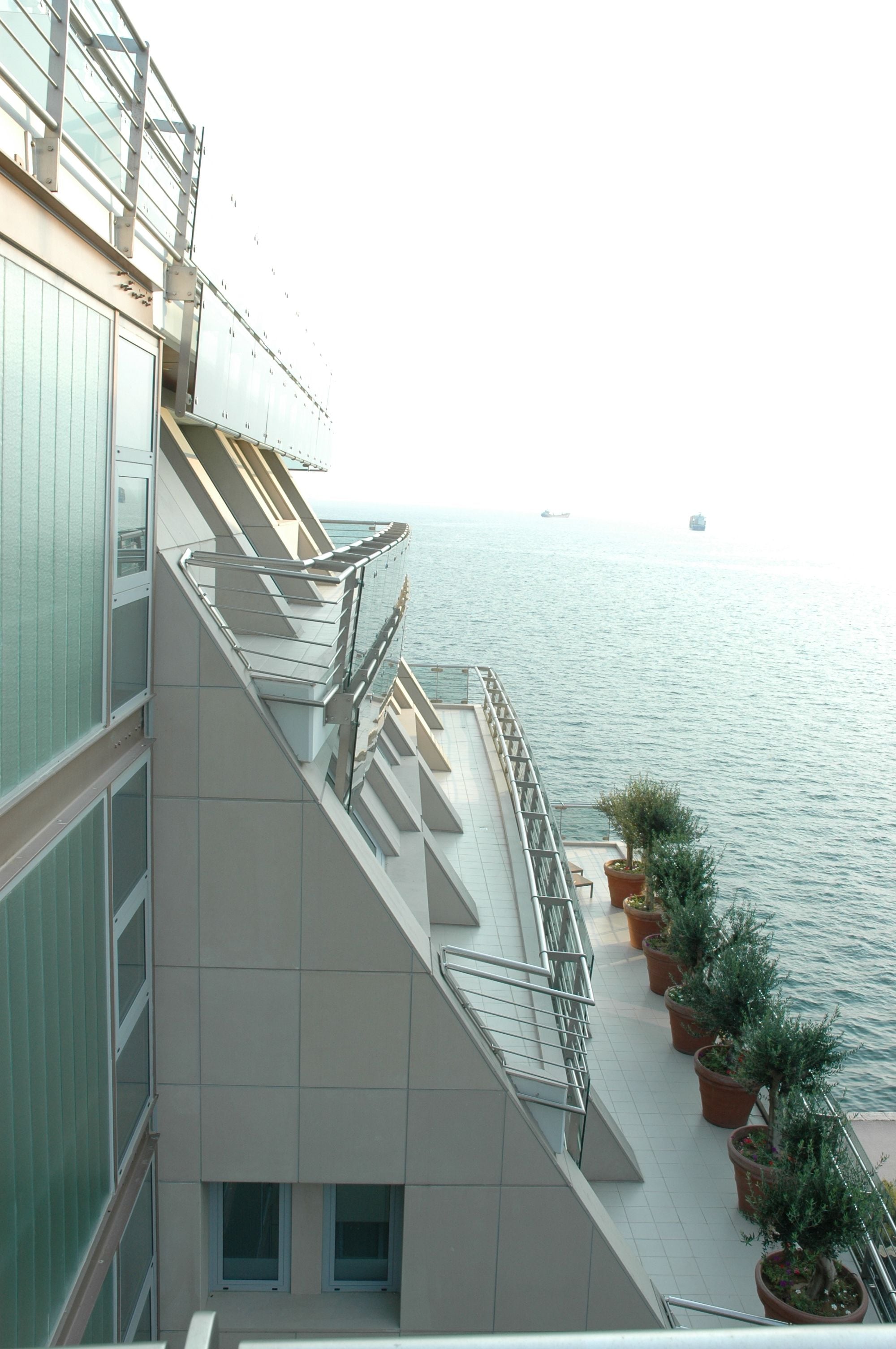 A balcony with a railing overlooking a body of water, potted plants, and a ship in the distance.