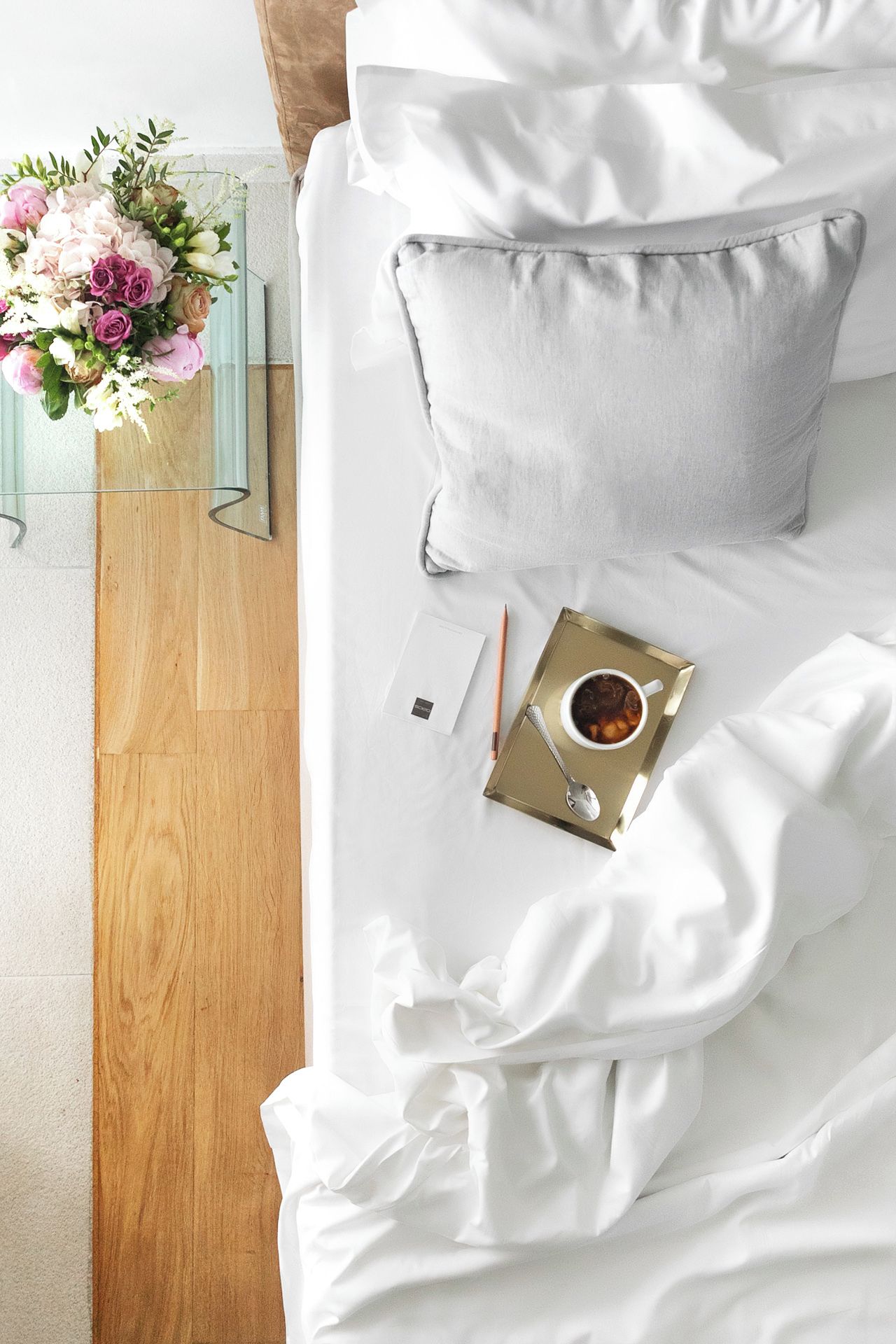 Una cama bien hecha con ropa blanca, una mesita de noche con una taza de té, un cuaderno y un lápiz.