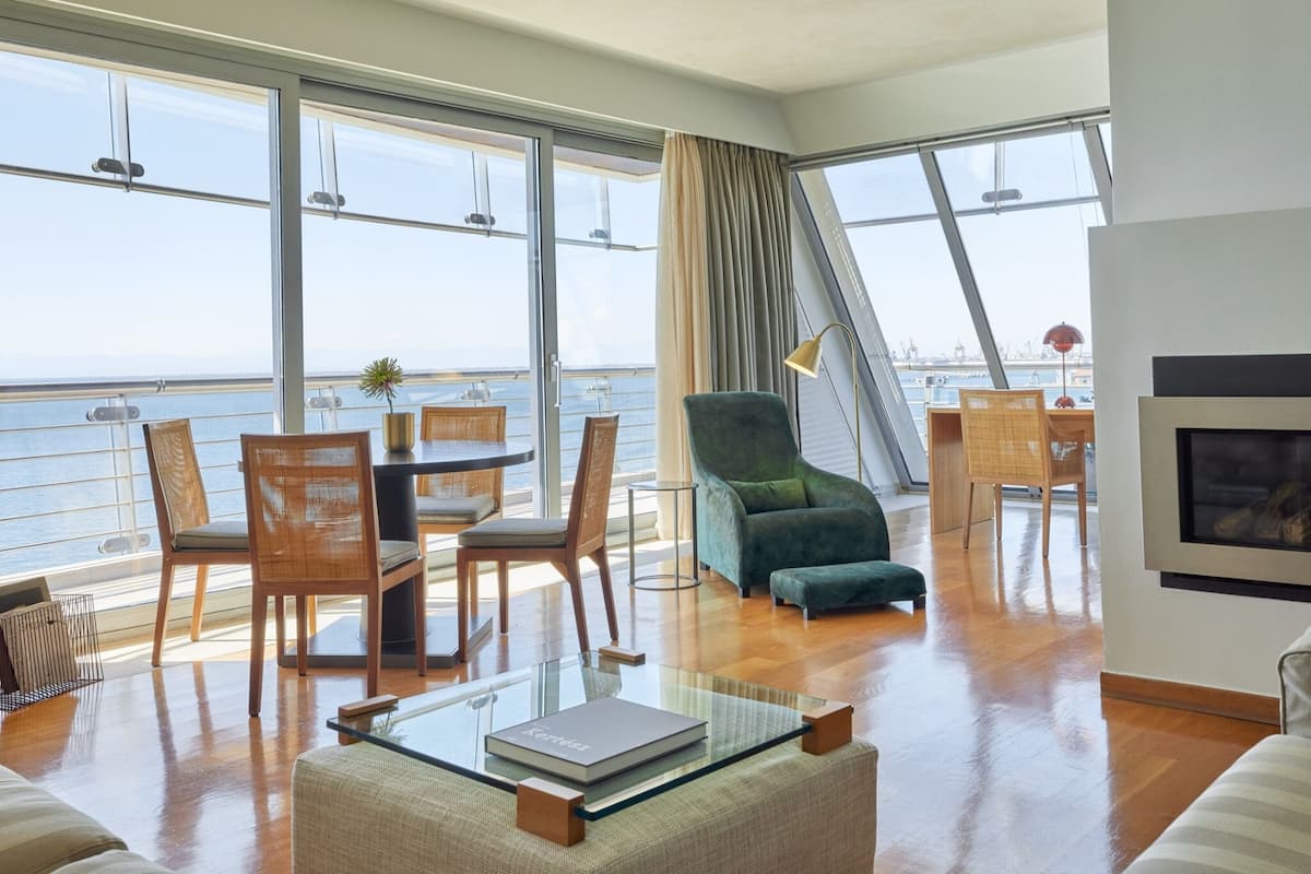 Een ruime zithoek met een groot raam met uitzicht op zee, een groene fauteuil, een glazen salontafel met een boek en een open haard.