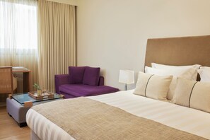 Habitación de hotel con cama, sofá, escritorio con lámpara y ventana con cortinas.