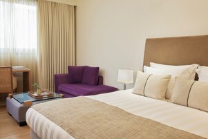 Habitación de hotel con cama, sofá, escritorio con lámpara y ventana con cortinas.