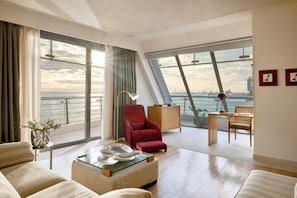 A modern living room with a sofa, armchair, and coffee table, a dining area with a chair and table, and large windows offering a view of the sea and cityscape.