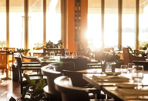 L’intérieur d’un restaurant, avec des tables dressées pour le service, des chaises et une grande fenêtre laissant entrer la lumière naturelle.
