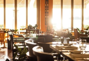 Interior de restaurante con mesas dispuestas para comer, sillas y un amplio ventanal que deja entrar luz natural.