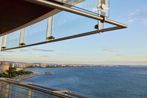 Un paisaje costero urbano con vista despejada al mar, una estructura imponente que recuerda a un castillo y un edificio moderno con fachada de vidrio.