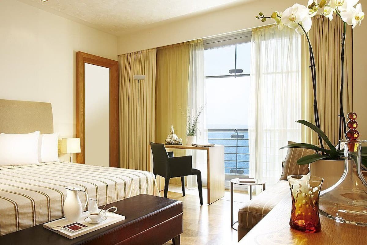 Een hotelkamer met een bed, een bureau, een stoel, een tafeltje, een vaas met bloemen en uitzicht op zee.