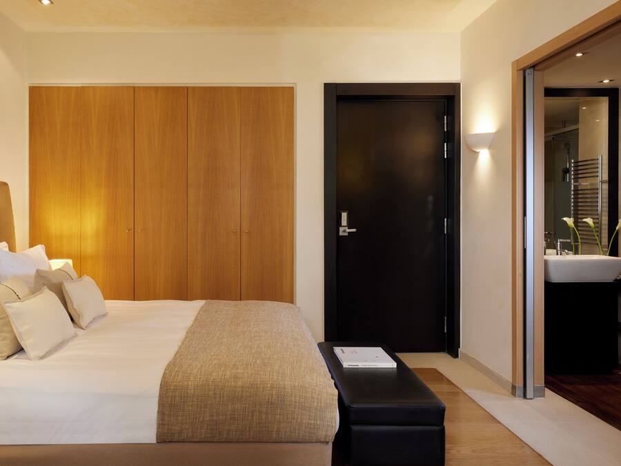 A modern hotel room with a large bed, a wooden wardrobe, a black door, and a bathroom with a sink and mirror.