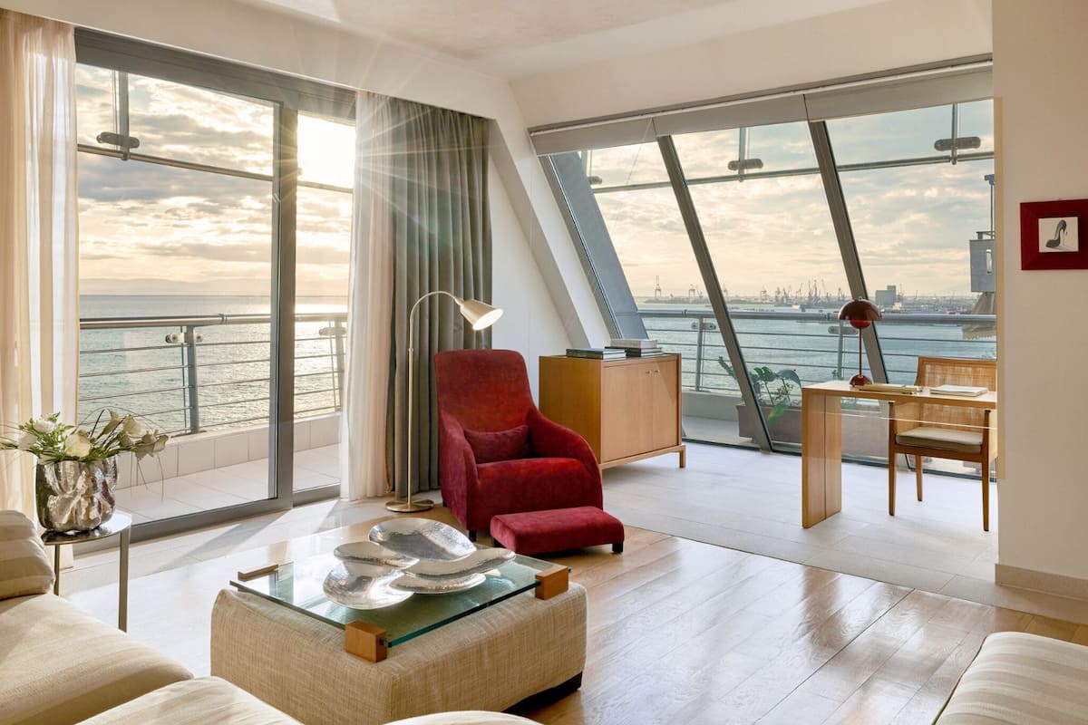 Een moderne woonkamer met een groot raam met uitzicht op een waterpartij, een rode fauteuil, een glazen salontafel en een houten bureau met stoel.