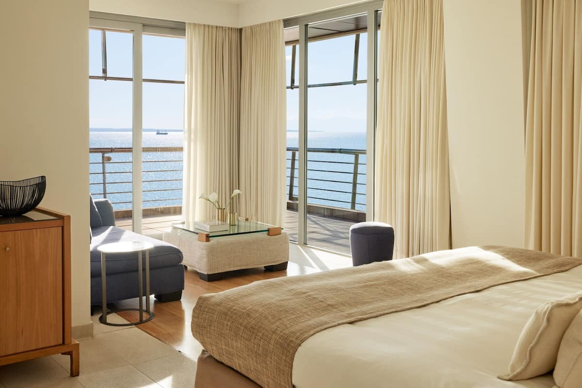 Een hotelkamer met een bed, een glazen tafel met een vaas, een stoel, een bank en uitzicht op de zee.
