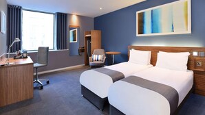 A hotel room with two beds, a desk, a chair, a round table, and a large abstract painting on the wall. - Travelodge Windsor Central (Windsor)