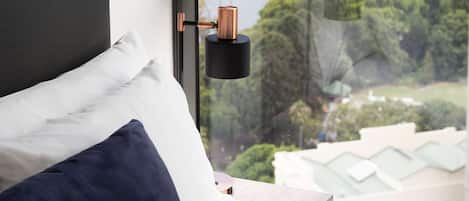 Una cama con almohadas blancas y azul marino, un cabecero negro y vistas de un paisaje urbano a través de una ventana con una lámpara de tono cobrizo.
