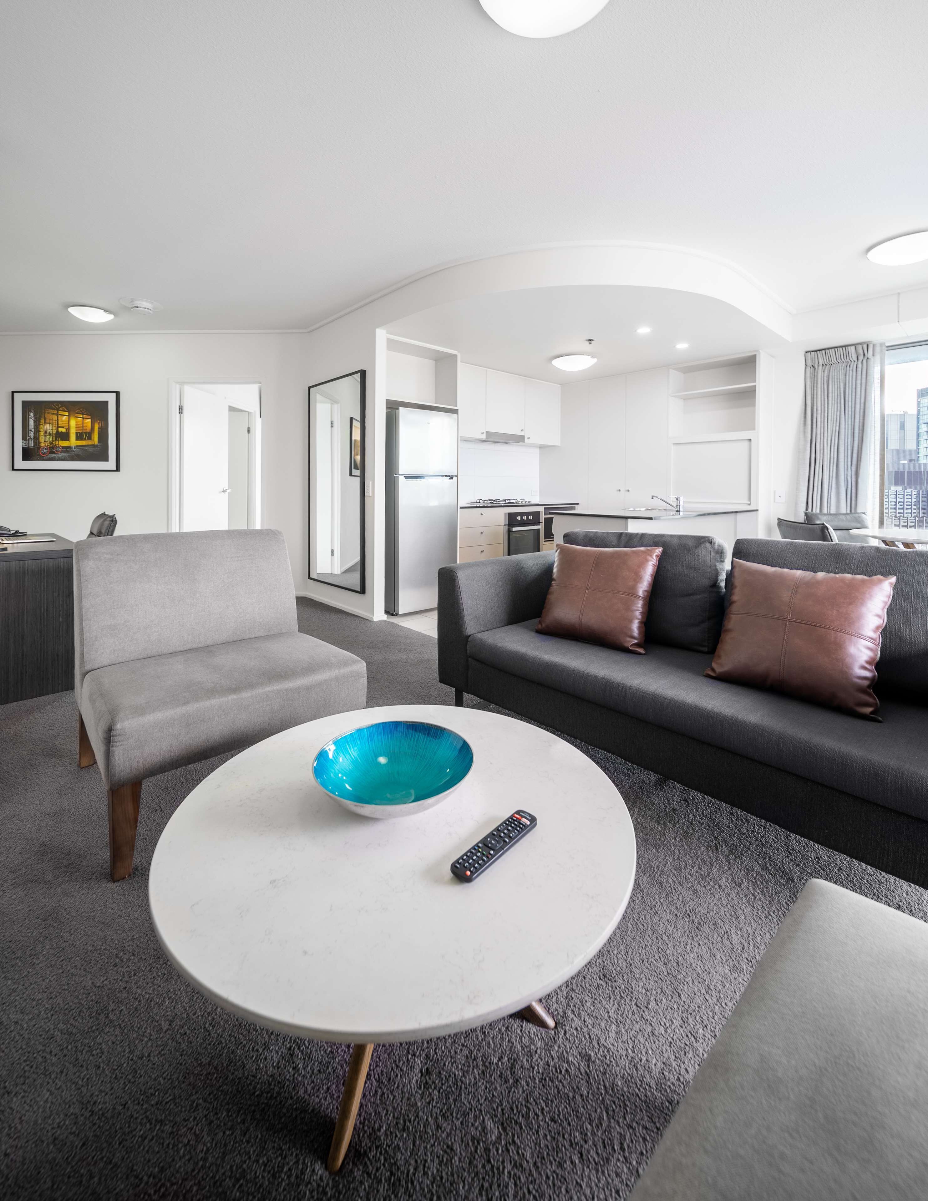 Een moderne woonkamer met een grijze bank, een ronde salontafel met een blauwe schaal, en een afstandsbediening.
