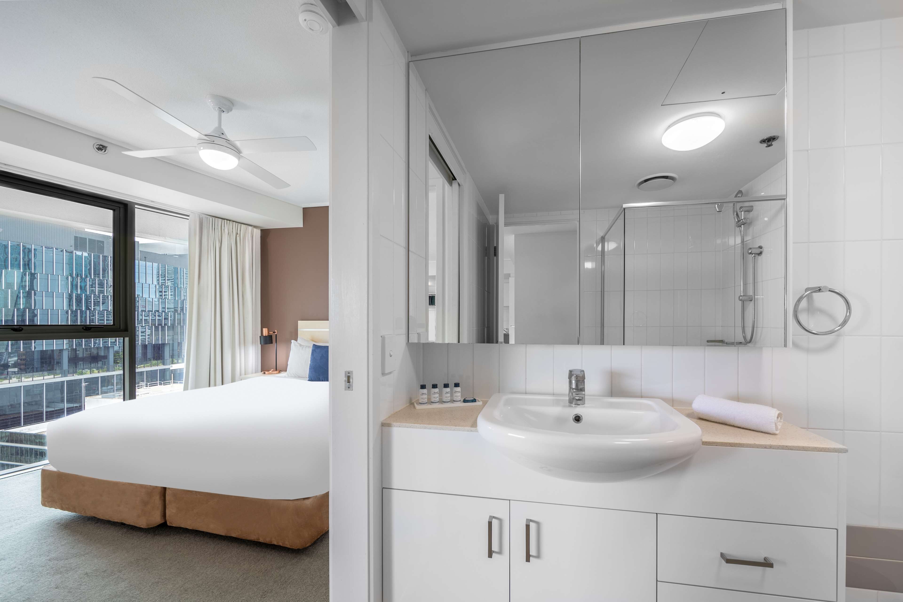 Una habitación de hotel moderna con una cama grande, un baño con lavamanos blanco y vistas a un paisaje urbano a través de un ventanal.