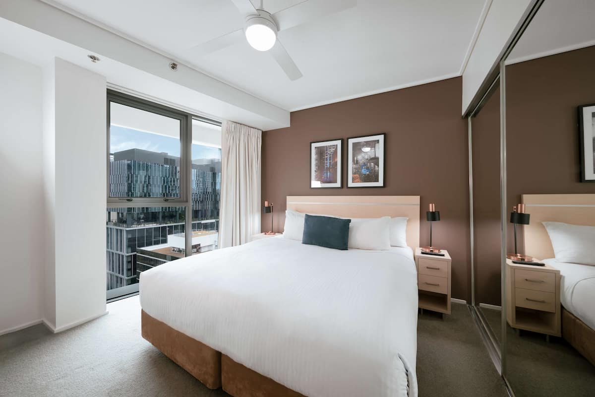 Ein modernes Hotelzimmer mit einem großen Bett, Nachttischen, einem Deckenventilator und Blick auf die Stadt durch ein großes Fenster.