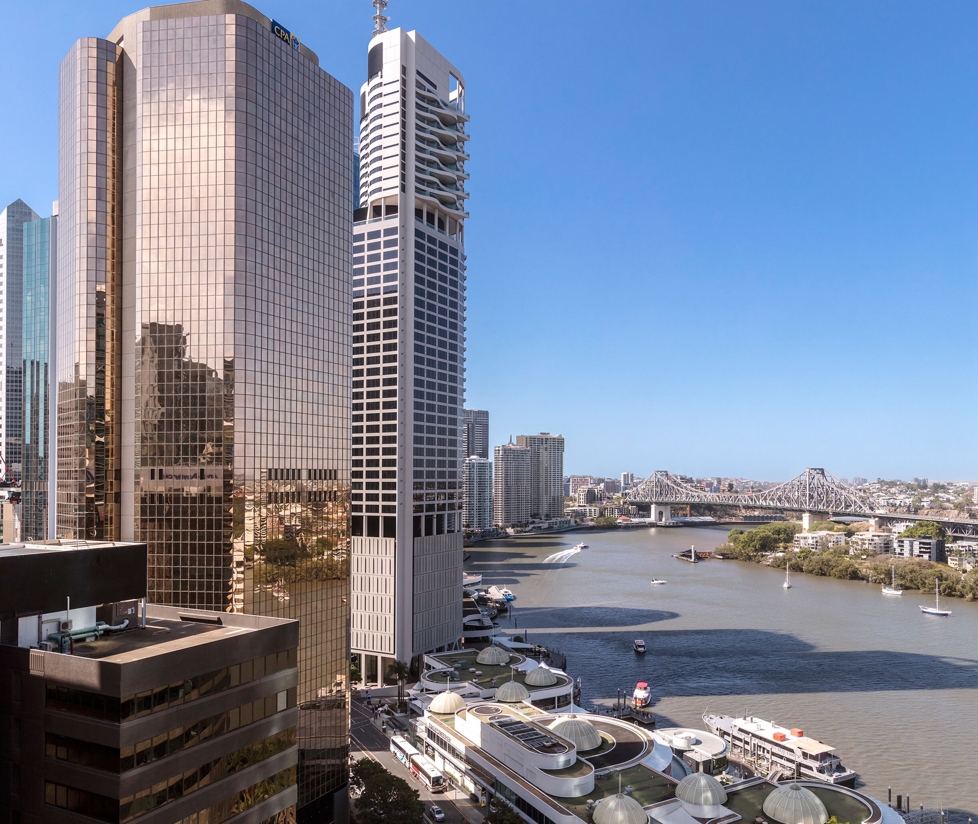 A cityscape with modern high-rise buildings, a river with boats, and a bridge in the distance.