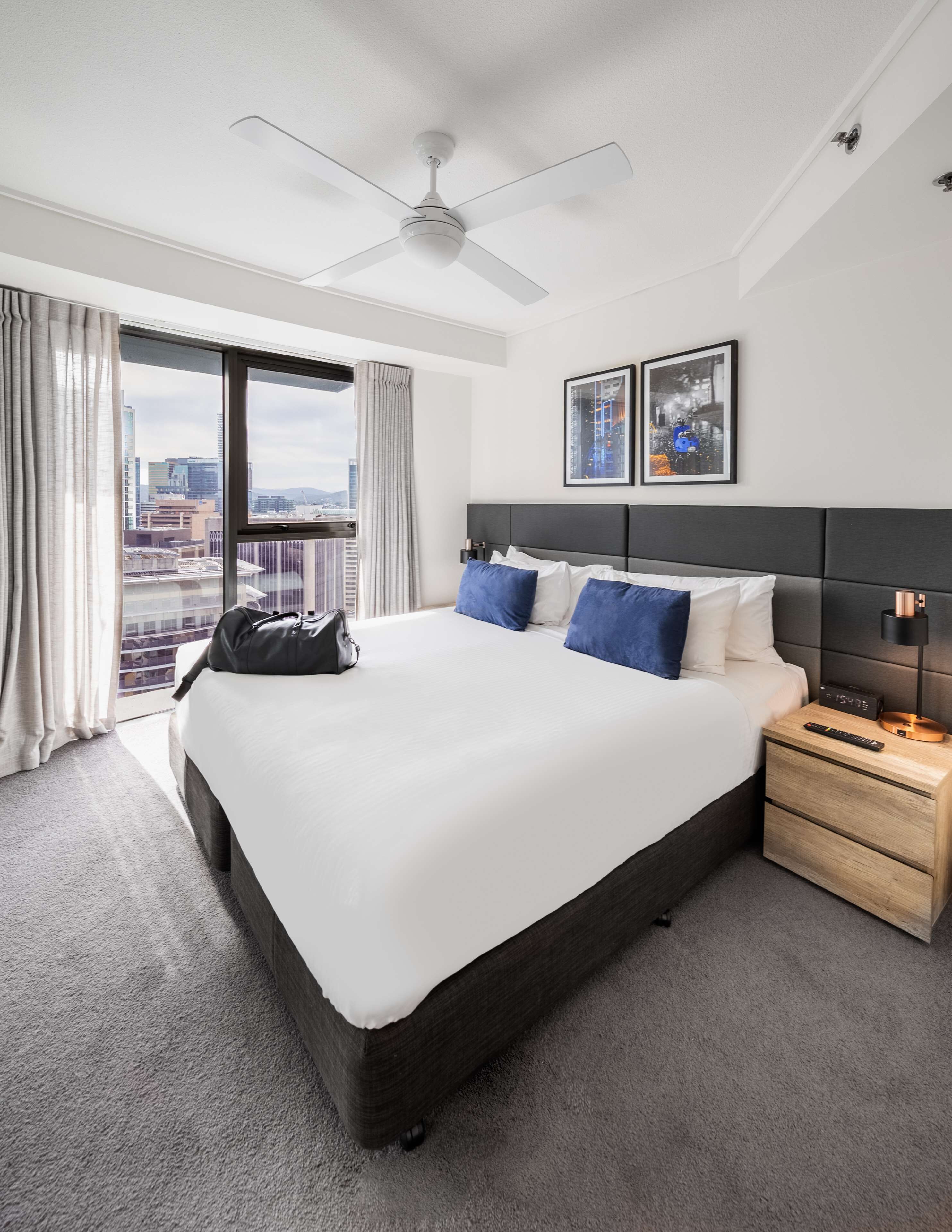 Une chambre d’hôtel moderne avec un grand lit, un ventilateur de plafond, une table de chevet et une vue sur la ville par la fenêtre.