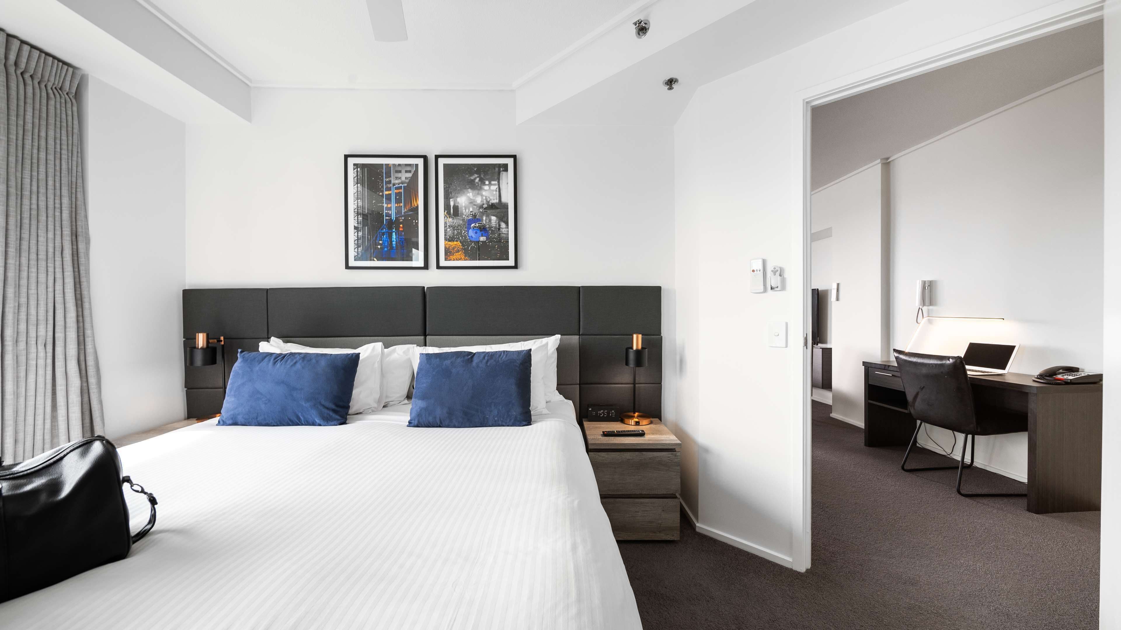 Una habitación de hotel moderna con una cama grande, un escritorio y una silla. Hay dos cuadros en la pared, una mesita de noche con una lámpara y un bolso negro sobre la cama.