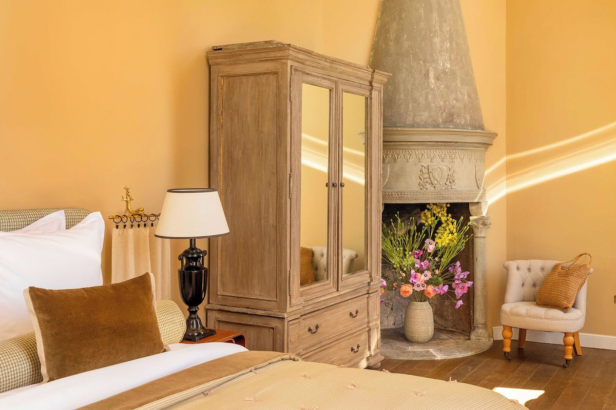 A room with a bed, a wooden cabinet, a fireplace with flowers, a chair, and a desk with a lamp.