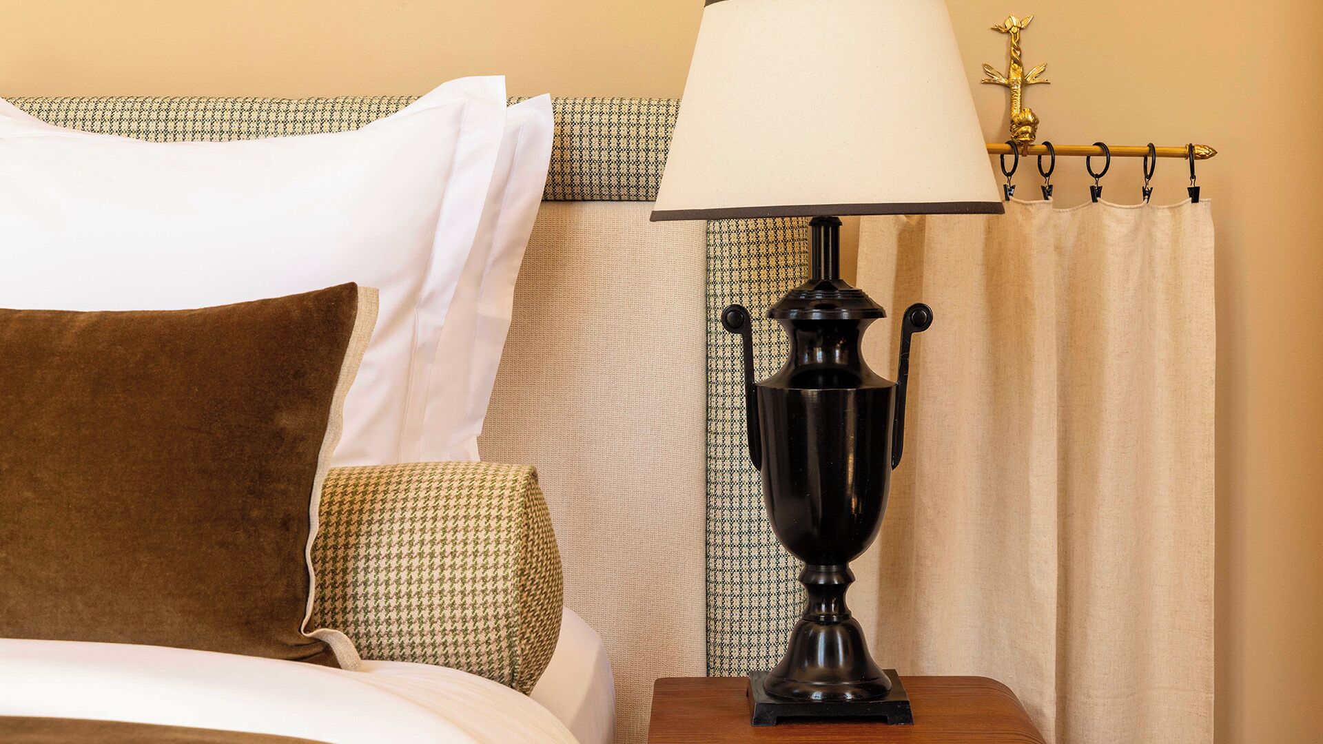 Una cama bien hecha con almohadas blancas y marrones, una manta estampada, una lámpara negra sobre una mesita de noche de madera y una cortina con una barra dorada.